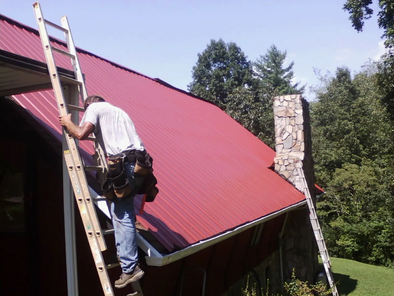 Roofer on a ladder beside a red metal roof and stone chimney — Soffit & Fascia Repair by Roofing Brothers in Stirling City